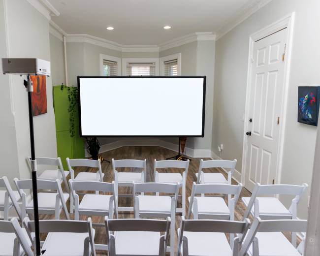 The image shows a room set up with white folding chairs facing a large blank screen, with a projector on a stand nearby.