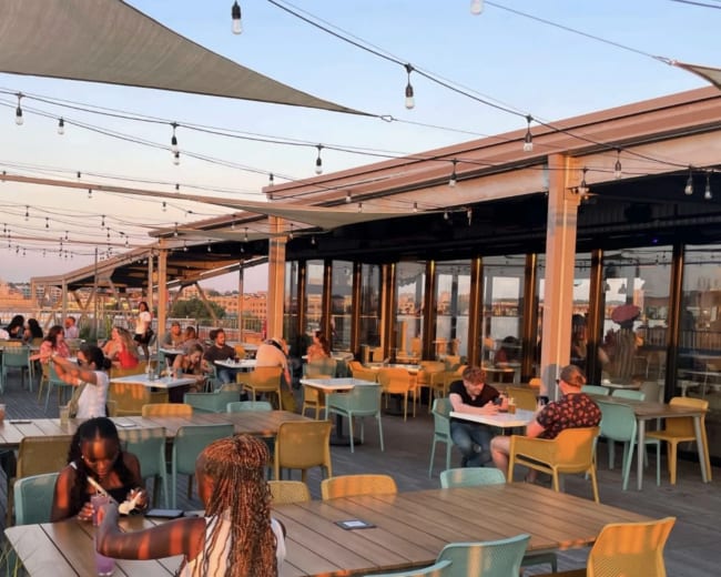 A rooftop restaurant with patrons dining at colorful tables under string lights during sunset.
