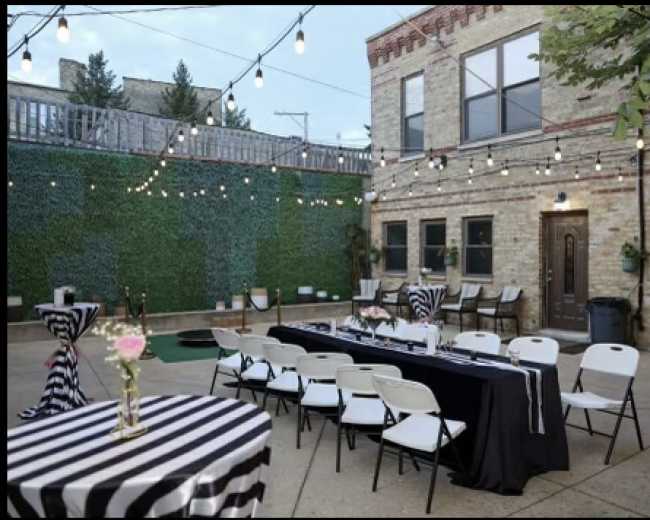 The image shows an outdoor event space with striped tablecloths, a long black table, chairs, and decorative string lights above, surrounded by greenery.