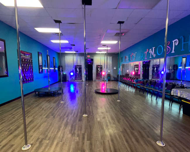 A dance studio featuring several poles, wooden flooring, and colorful lights, with a backdrop of blue walls and mirrors.