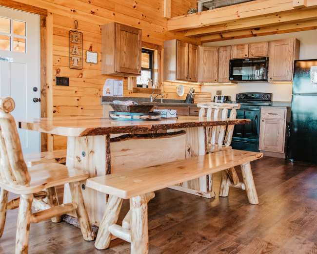 The image shows a wooden kitchen with a table and benches made of light-colored wood, surrounded by rustic cabinetry and appliances.