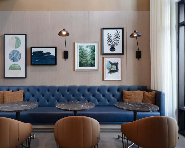 The image shows a modern lounge area with a blue tufted bench, marble-topped tables, and framed artwork on the walls.