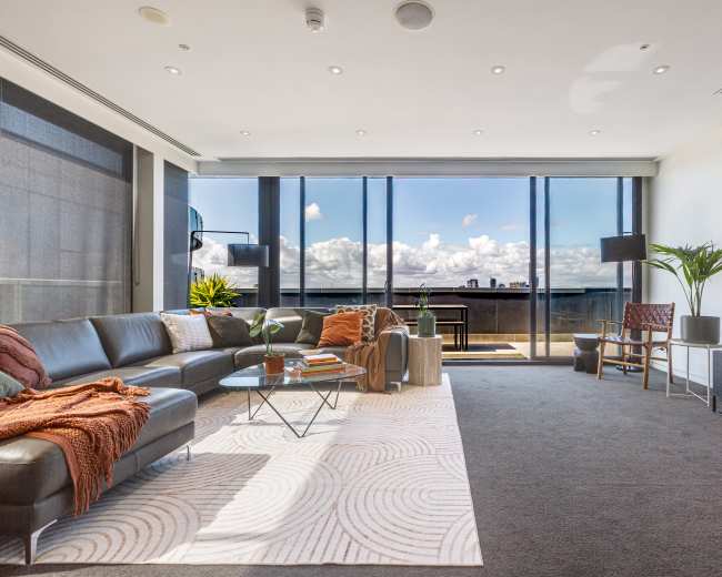The image depicts a modern living room with a large sectional sofa, a coffee table, and a view of a balcony overlooking a bright sky with clouds.