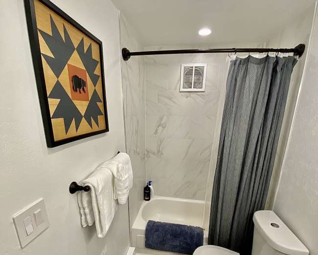 The image shows a compact bathroom featuring a bathtub with a shower curtain, a toilet, a sink with a dark countertop, and wall art above the tub.