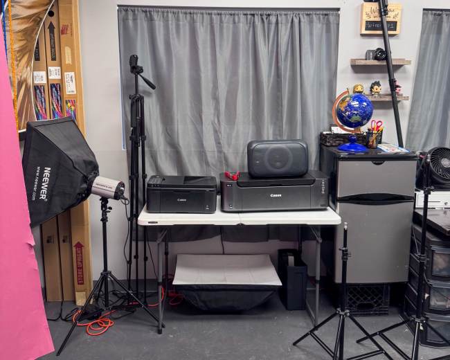 The image shows a workspace featuring two printers, a monitor, a small fridge, and various photographic equipment against a backdrop of gray curtains and organized shelves.