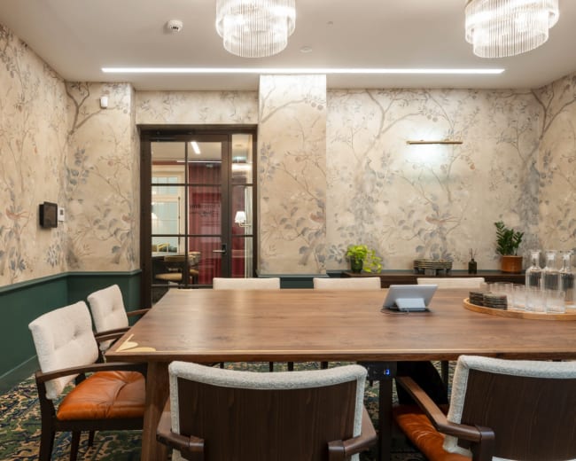 The image shows a conference room with a large wooden table surrounded by upholstered chairs, adorned with floral wallpaper and equipped with a glass door leading to another room.