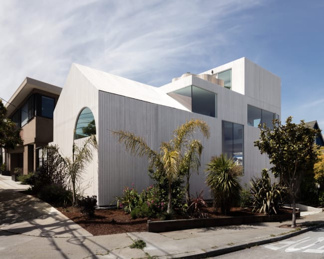 A modern, angular house with a white wood exterior and large windows is located on a sloped street, surrounded by landscaped greenery and other residential buildings.