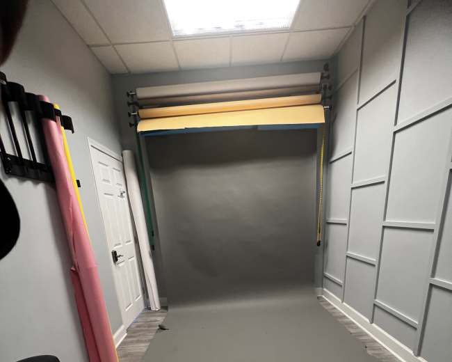 A photography studio with a gray backdrop set up against a wall, flanked by colorful paper rolls and a chair in the corner.