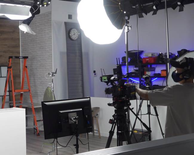 A woman stands behind a white desk in a studio set up for a television broadcast, with cameras and lighting equipment arranged around her.