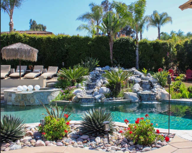 The image shows a landscaped backyard with a swimming pool, a small waterfall, tropical plants, and lounge chairs.