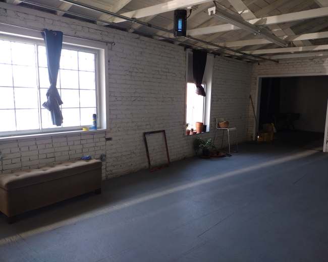 A spacious room with white brick walls, two large windows with curtains, a bench, and various plants along with a ladder and other furnishings in the background.