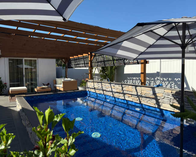 A well-lit backyard features a swimming pool with a stone border, surrounded by lounging chairs and shade umbrellas under a wooden pergola.