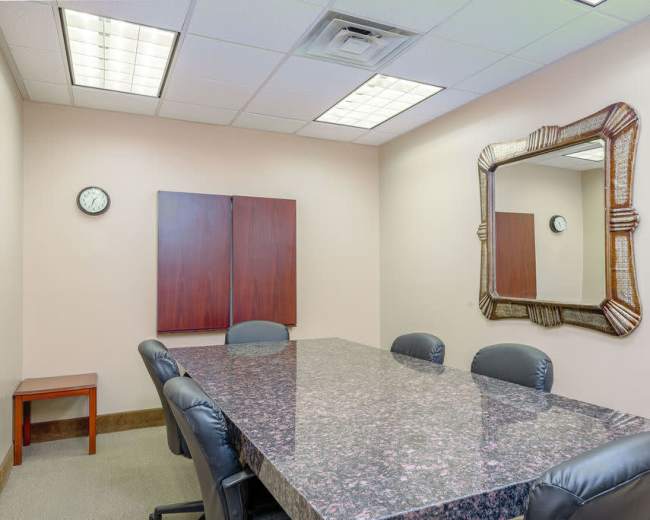 A conference room features a dark marble table surrounded by black chairs, with a clock on the wall and a large decorative mirror.