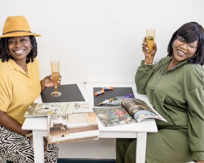 Two women sit at a table, each holding a drink, surrounded by magazines and crafting supplies.