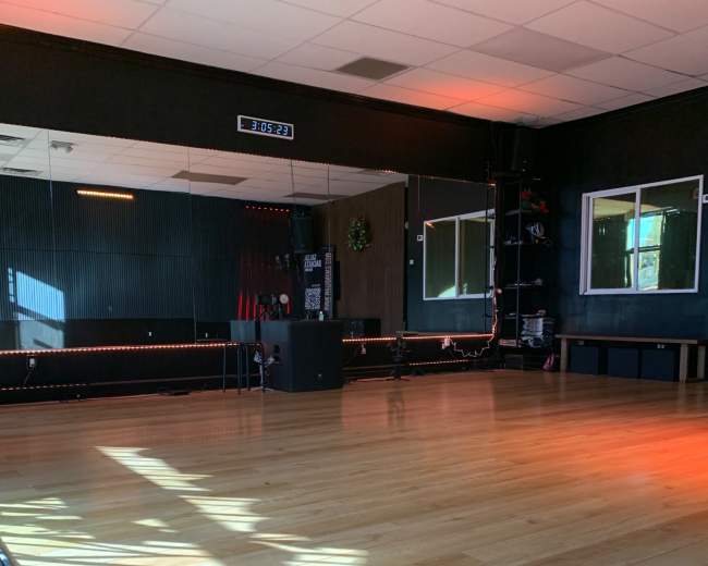 The image shows an empty fitness studio with wooden flooring, a large mirror, and windows illuminated by sunlight.