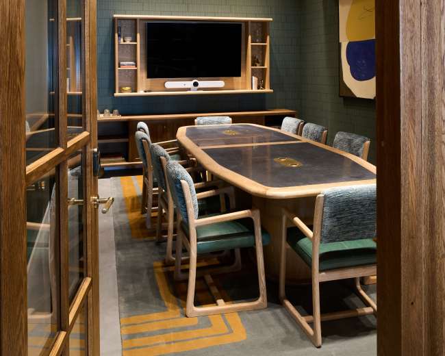 A modern conference room features a large wooden table surrounded by upholstered chairs, with a television mounted on the wall and a decorative carpet.
