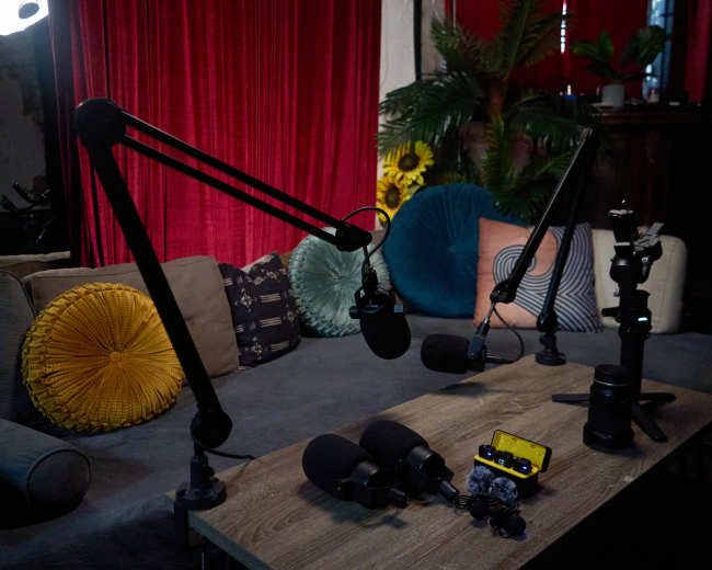 The image shows a podcast setup with multiple microphone stands, a wooden table, and a colorful couch adorned with pillows.