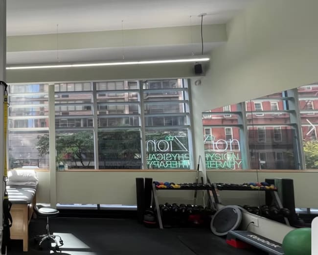 The image shows a bright therapy room with large windows, a row of therapy tables, and exercise equipment arranged along the wall.