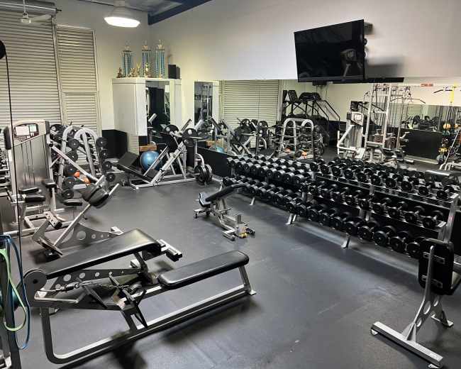 The image shows a gym interior with various workout equipment, including weight benches, dumbbells, and a television mounted on the wall.