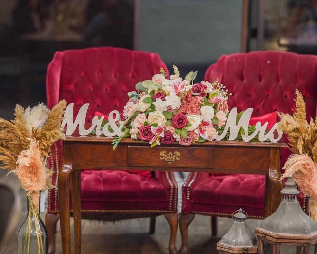The image shows a cozy seating area with two red velvet chairs, a wooden table adorned with a flower bouquet, and decorative lanterns.