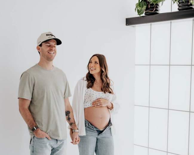 A couple stands together in a bright studio, with one partner holding their pregnant belly while smiling at the camera.