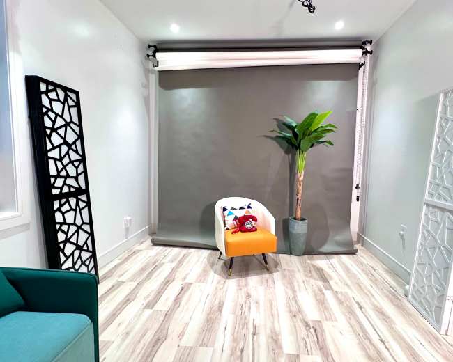 The image shows a minimalist room with a gray backdrop, a bright orange chair, a potted plant, and decorative screens on either side.