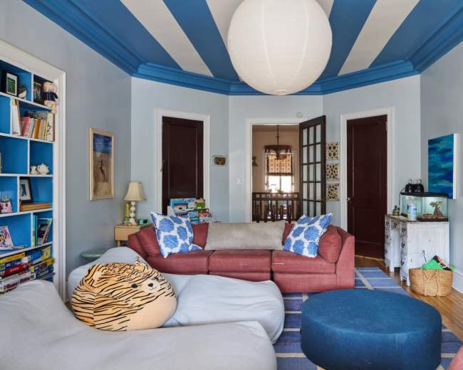 A cozy living room features a sectional sofa, patterned cushions, a large bean bag, and a circular rug, with blue-striped ceilings and a lantern-style light fixture.