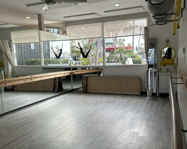 The image shows a light-filled room with large windows, a ballet barre along one wall, and mirrors reflecting the space, set up for fitness or dance activities.