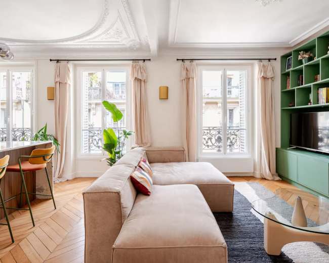 The image shows a well-lit living room featuring a beige sectional sofa, a green bookshelf, and large windows with beige curtains.