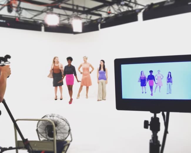 A group of five models stands in front of a white backdrop while being filmed by a camera on a tripod.