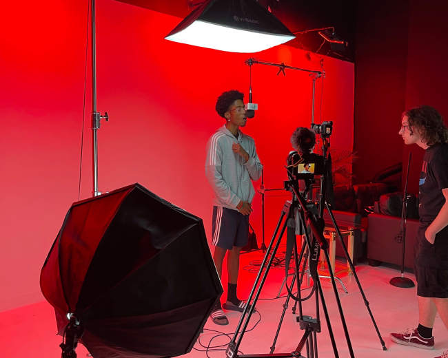A young man in a gray sweatshirt standing in a studio with red lighting, surrounded by camera equipment and two other individuals.