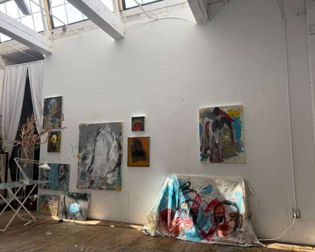 The image shows an art studio with a white wall displaying several abstract paintings and a draped table with colorful artwork.