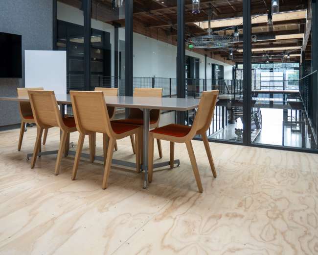 A modern conference room features a large wooden table surrounded by six wooden chairs on a plywood floor.