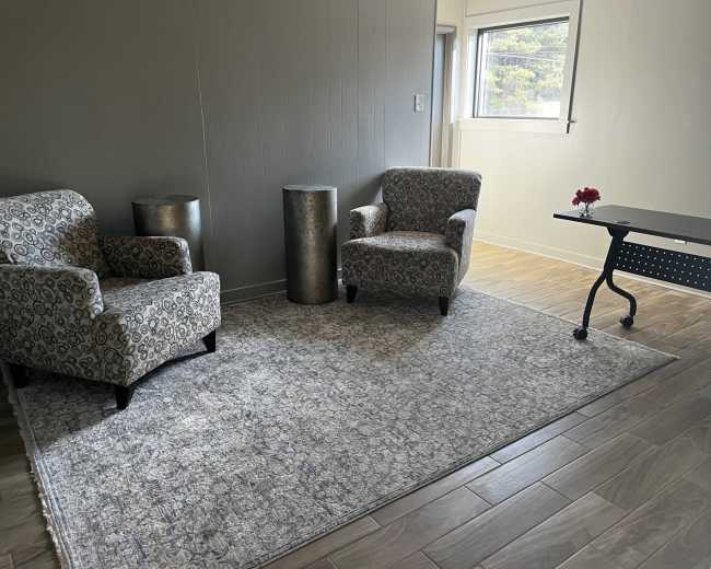 The image shows a small, modern room with two patterned chairs, a metallic cylindrical table, and a small desk on a wooden floor.