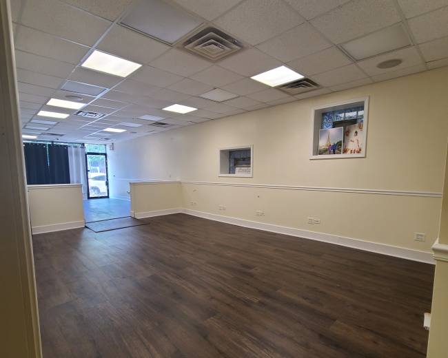 The image shows a vacant interior space with wooden floors, white walls, and a large window at one end.