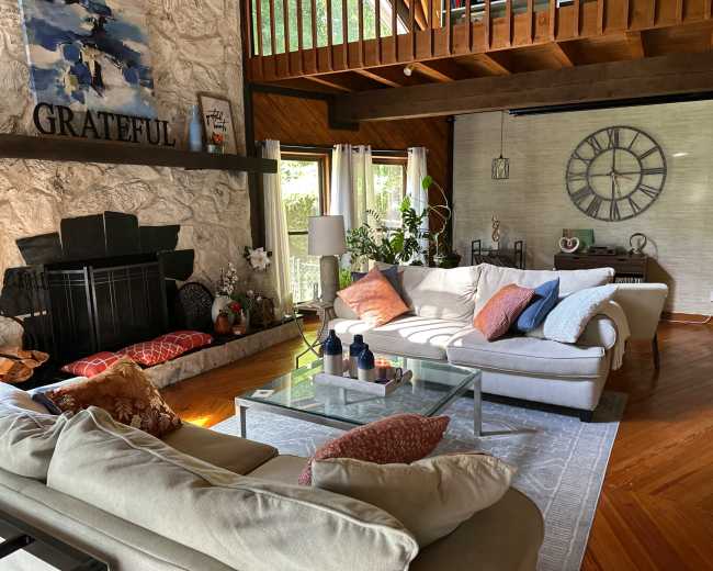 The image depicts a spacious living room featuring two sofas, a coffee table, a stone fireplace, and a large clock on the wall.
