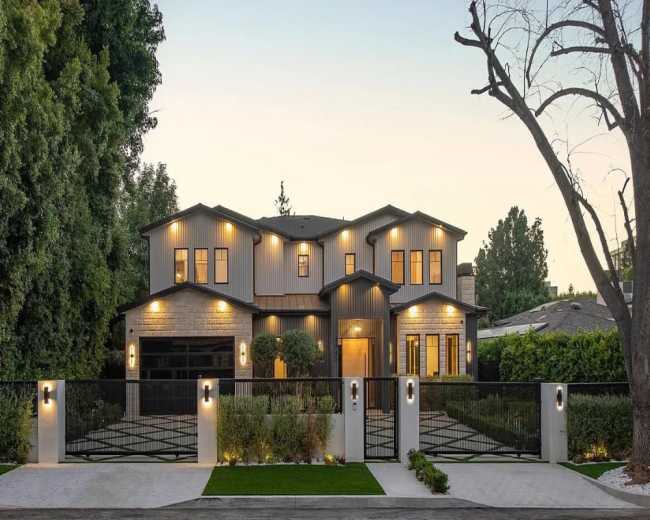 The image shows a modern two-story house with a mix of stone and metal siding, surrounded by greenery and featuring a gated entrance with landscaped pathways.