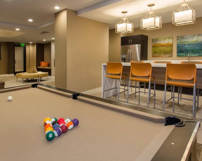 The image shows a recreational room featuring a pool table with a set of billiard balls and a bar area with seating and artwork on the wall.