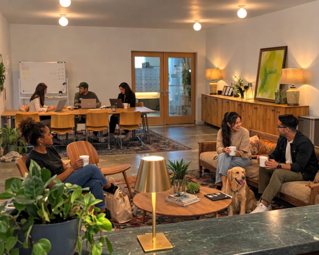 A cozy shared workspace features people engaged in conversation and working at tables, with plants and a dog present in the inviting environment.