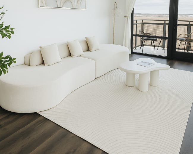 The image shows a modern living room with a curved white sofa, a round coffee table, and large windows that open to a balcony view.