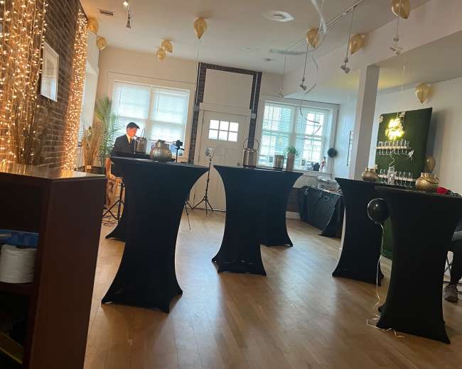 A spacious indoor event space with dark tables set up, string lights on the walls, and balloons hanging from the ceiling.