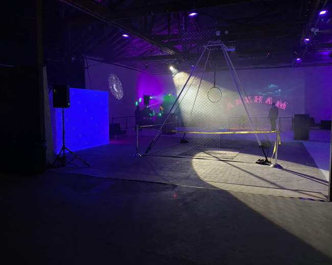 The image shows an industrial space set up for an event, featuring a large mesh structure with a hoop, a blue illuminated backdrop, and various lights in the background.
