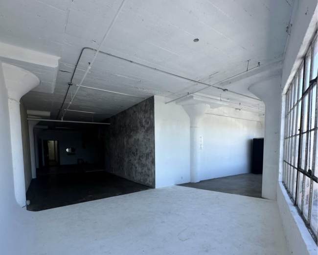 The image shows a spacious, partially empty indoor area with white walls, a concrete floor, and large windows allowing natural light.