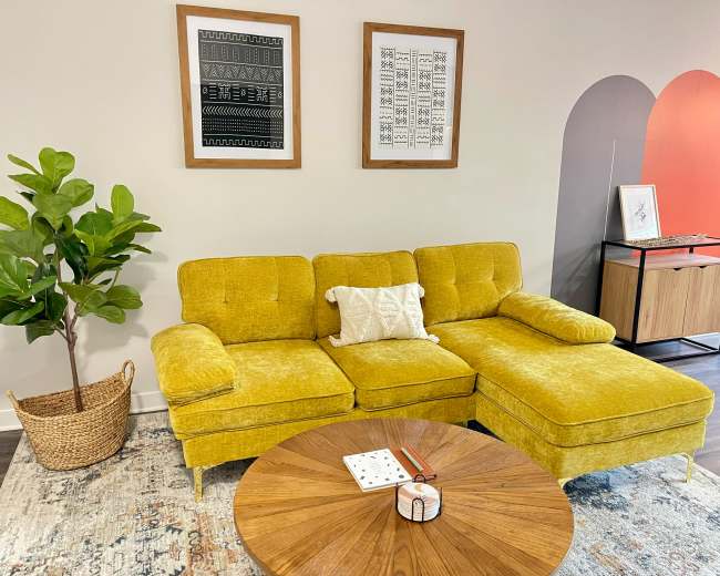 A yellow sectional sofa with a decorative pillow sits in a modern room, accompanied by a round wooden coffee table and a potted plant.