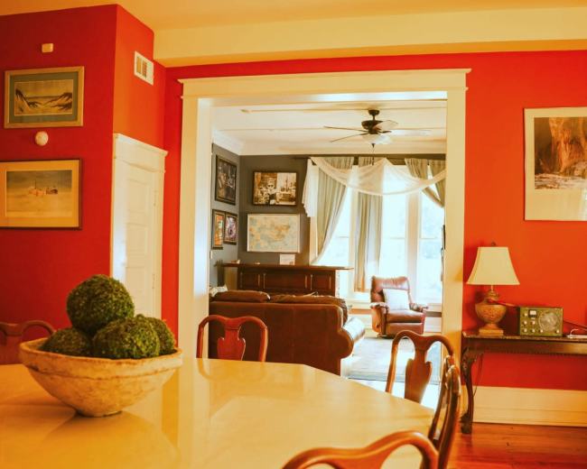 The image shows a dining room with a round table and a chandelier, leading into a cozy living room adorned with framed pictures and a large window.