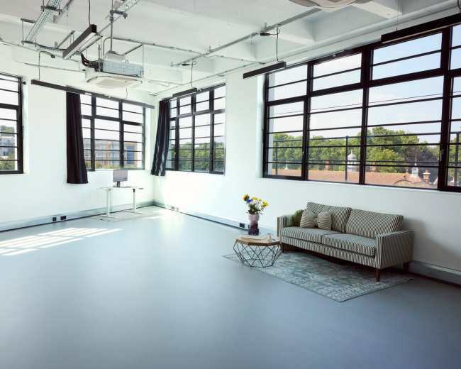 The image shows a bright, spacious room with large windows, a couch, a small table with flowers, and a desk against the wall.
