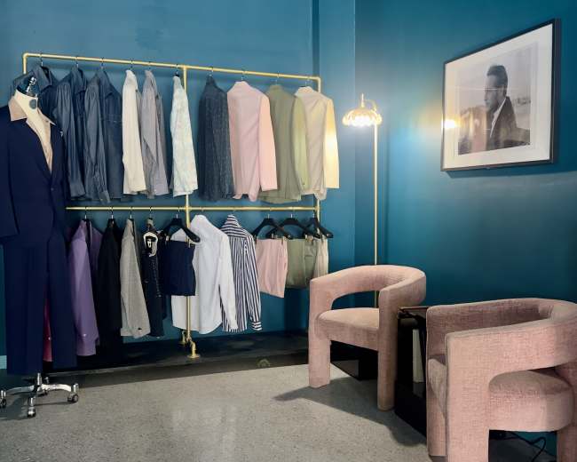 The image shows a clothing display with various shirts and jackets hanging on a golden rack against a teal wall, accompanied by two pink chairs and a framed black-and-white photograph.