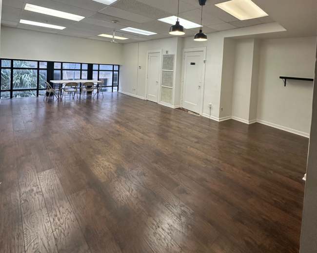 The image shows a spacious, well-lit room with hardwood flooring, large windows, and a dining table set for several people.