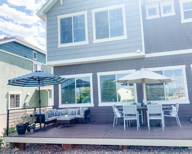 The image shows a house with a large deck featuring a dining table set under an umbrella and a seating area with a couch and an umbrella.