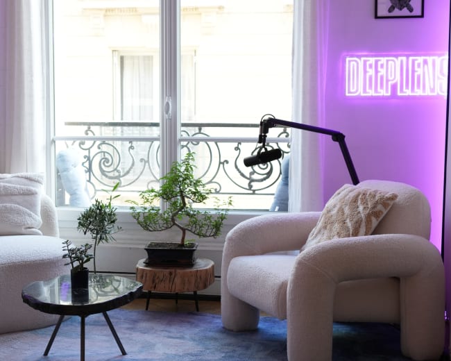 The image shows a modern living room with a white curved sofa and chair, a circular table, and large windows framed by sheer curtains, all illuminated by purple lighting and a neon sign.